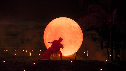 A dramatic scene with two people dancing in front of a large glowing sphere resembling a sun or moon. The environment is dark, with silhouettes of palm trees and several small illuminated lights scattered around, adding to the mystical atmosphere.
