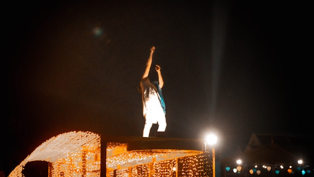 A person is standing on a platform adorned with festive lights, raising an arm as if engaging with an audience. The scene is lit by various light sources, creating a vibrant and dynamic atmosphere against a dark background.