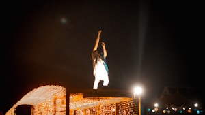 A person is standing on a platform adorned with festive lights, raising an arm as if engaging with an audience. The scene is lit by various light sources, creating a vibrant and dynamic atmosphere against a dark background.