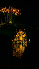 A group of people appears to be performing in a body of water under soft, warm light. They are gathered beneath a large, artistic tree-like structure adorned with illuminated round objects, which resemble lanterns or orbs. The reflections in the water add a vibrant glow to the scene, enhancing the mysterious and enchanting atmosphere.