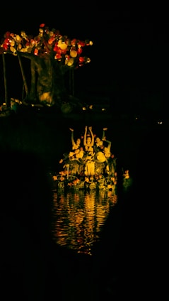 A group of people appears to be performing in a body of water under soft, warm light. They are gathered beneath a large, artistic tree-like structure adorned with illuminated round objects, which resemble lanterns or orbs. The reflections in the water add a vibrant glow to the scene, enhancing the mysterious and enchanting atmosphere.
