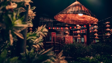 A nighttime scene featuring traditional lanterns hanging above a lush garden area. Wooden structures and greenery create a tranquil setting, with warm, glowing lights creating a cozy atmosphere. The backdrop includes a building with detailed decorations, contributing to the overall ambiance.