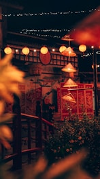 An illuminated scene at night featuring traditional red lanterns hanging along a building decorated with intricate patterns. Strings of small lights and a row of conical straw hats, also lit, add to the ambiance. The foreground includes lush greenery and a wooden railing.