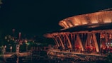 A vibrant, warmly lit restaurant with a unique circular structure made of wood, featuring open seating and patrons dining. The ambiance is serene with lanterns providing additional light, and a reflection can be seen on a water body nearby.