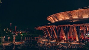 A vibrant, warmly lit restaurant with a unique circular structure made of wood, featuring open seating and patrons dining. The ambiance is serene with lanterns providing additional light, and a reflection can be seen on a water body nearby.