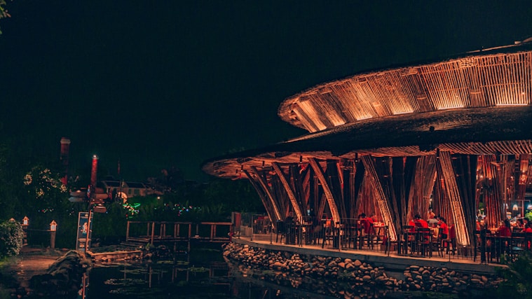A vibrant, warmly lit restaurant with a unique circular structure made of wood, featuring open seating and patrons dining. The ambiance is serene with lanterns providing additional light, and a reflection can be seen on a water body nearby.