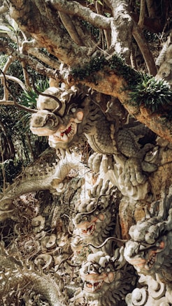 A detailed sculpture of multiple dragon heads emerges from a rocky and natural setting. The intricate carvings show fierce expressions with open mouths, sharp teeth, and long, curling horns. Each dragon head is surrounded by lush greenery, blending seamlessly with the tree branches above.