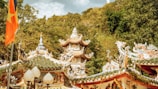 A traditional Asian temple with intricate architectural details, including ornate roofs with elaborate carvings and decorations. There is a flag with a yellow star on a red background prominently displayed on the left side. The temple is surrounded by lush green trees and a hillside in the background.