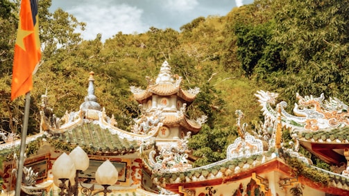 A traditional Asian temple with intricate architectural details, including ornate roofs with elaborate carvings and decorations. There is a flag with a yellow star on a red background prominently displayed on the left side. The temple is surrounded by lush green trees and a hillside in the background.