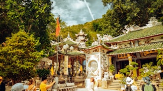 A vibrant scene in a traditional Asian temple complex surrounded by lush greenery. Intricate architectural details adorn the temple buildings, which are decorated with colorful patterns and sculptures. Several visitors, some wearing hats, are walking around the temple grounds. Flags and banners add to the cultural richness of the setting.