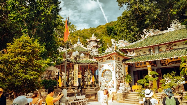 A vibrant scene in a traditional Asian temple complex surrounded by lush greenery. Intricate architectural details adorn the temple buildings, which are decorated with colorful patterns and sculptures. Several visitors, some wearing hats, are walking around the temple grounds. Flags and banners add to the cultural richness of the setting.