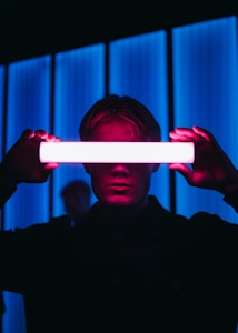 A person holds a bright, glowing light bar horizontally in front of their face, casting a neon pink and blue hue. The background features vertical blue light strips, creating a futuristic atmosphere.