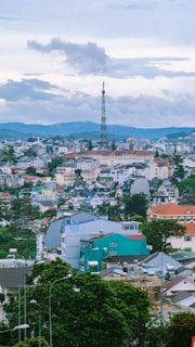 A scenic cityscape featuring a prominent radio tower surrounded by a densely packed area of colorful buildings and houses. The landscape is set against a background of rolling hills and a partly cloudy sky, creating a blend of urban and natural elements.