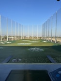 A modern driving range with multiple circular target areas on a well-maintained grass field. Tall netting surrounds the area, extending upwards to contain golf balls. The sky is clear and blue, and there are distant trees and buildings visible in the background.