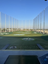 A modern driving range with multiple circular target areas on a well-maintained grass field. Tall netting surrounds the area, extending upwards to contain golf balls. The sky is clear and blue, and there are distant trees and buildings visible in the background.