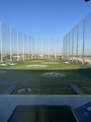 A modern driving range with multiple circular target areas on a well-maintained grass field. Tall netting surrounds the area, extending upwards to contain golf balls. The sky is clear and blue, and there are distant trees and buildings visible in the background.