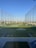 A modern driving range with multiple circular target areas on a well-maintained grass field. Tall netting surrounds the area, extending upwards to contain golf balls. The sky is clear and blue, and there are distant trees and buildings visible in the background.