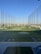 A modern driving range with multiple circular target areas on a well-maintained grass field. Tall netting surrounds the area, extending upwards to contain golf balls. The sky is clear and blue, and there are distant trees and buildings visible in the background.