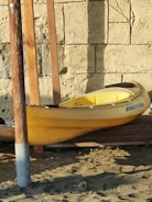 A yellow kayak rests on a wooden platform against a textured stone wall. The kayak is upright, with its bow slightly elevated. Two wooden posts are positioned vertically beside the kayak, and the ground is covered with sand and small plants.