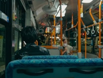 The interior of a city bus with passengers seated. One person can be seen looking out the window while using a mobile phone, and another person is wearing a face mask and may be interacting with a mobile phone. The bus is equipped with overhead handrails and straps for standing passengers.