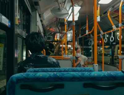 The interior of a city bus with passengers seated. One person can be seen looking out the window while using a mobile phone, and another person is wearing a face mask and may be interacting with a mobile phone. The bus is equipped with overhead handrails and straps for standing passengers.