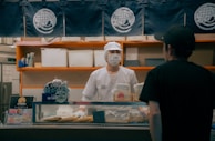 A person wearing a white uniform and a face mask, standing behind a shop counter. The setting is a food establishment with signage and displays, including packages and a price sign. Another person wearing a black shirt and cap is standing in front of the counter.
