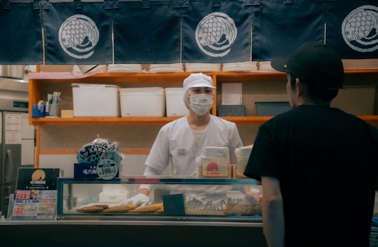 A person wearing a white uniform and a face mask, standing behind a shop counter. The setting is a food establishment with signage and displays, including packages and a price sign. Another person wearing a black shirt and cap is standing in front of the counter.