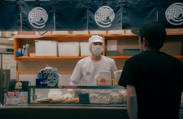 A person wearing a white uniform and a face mask, standing behind a shop counter. The setting is a food establishment with signage and displays, including packages and a price sign. Another person wearing a black shirt and cap is standing in front of the counter.