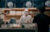 A person wearing a white uniform and a face mask, standing behind a shop counter. The setting is a food establishment with signage and displays, including packages and a price sign. Another person wearing a black shirt and cap is standing in front of the counter.