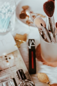 A variety of makeup brushes are neatly arranged in a holder, accompanied by several cosmetics items including a lipstick and perfume. The setting has an elegant yet casual feel, with soft lighting highlighting the textures and colors of the beauty products.