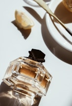 A bottle of perfume with a geometric design sits on a white surface, surrounded by a slice of lemon and a plate with a butter spreader. The sunlight creates soft shadows and highlights, adding a warm glow to the scene.