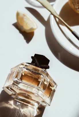 A bottle of perfume with a geometric design sits on a white surface, surrounded by a slice of lemon and a plate with a butter spreader. The sunlight creates soft shadows and highlights, adding a warm glow to the scene.