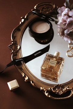 A vintage-style gold ornate mirror reflects a table arrangement. On the mirror, there is a bottle of Chanel perfume, a cream cosmetic container, and a black ribbon with the Chanel logo. In the background, a bouquet of pink flowers and a small white cube with a logo are visible. The setting conveys an elegant and luxurious aesthetic.