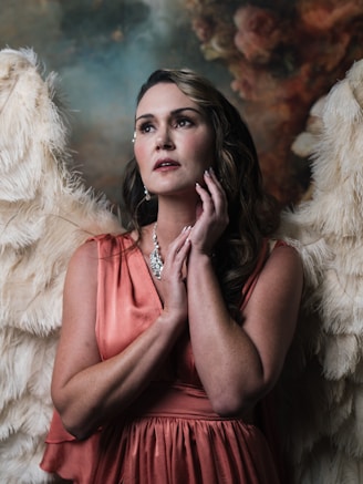 A person is posed at the center, dressed in a flowing coral dress and adorned with a sparkling necklace and earrings. Angelic, fluffy wings are attached to their back, creating a serene, celestial atmosphere. The background is an abstract blend of dark and light tones, suggesting a soft, heavenly scene.
