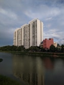 A tall, modern apartment building with multiple floors stands prominently near a body of water. The building is white with a sleek design and numerous balconies. In front of it, a smaller, reddish building is visible, along with lush green trees lining the water. The water reflects the apartment building and surrounding foliage. The sky is expansive and partly cloudy, with a serene blue hue.