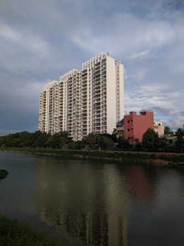 A tall, modern apartment building with multiple floors stands prominently near a body of water. The building is white with a sleek design and numerous balconies. In front of it, a smaller, reddish building is visible, along with lush green trees lining the water. The water reflects the apartment building and surrounding foliage. The sky is expansive and partly cloudy, with a serene blue hue.