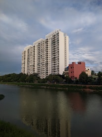 A tall, modern apartment building with multiple floors stands prominently near a body of water. The building is white with a sleek design and numerous balconies. In front of it, a smaller, reddish building is visible, along with lush green trees lining the water. The water reflects the apartment building and surrounding foliage. The sky is expansive and partly cloudy, with a serene blue hue.