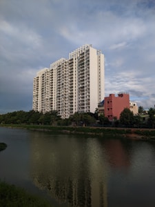 A tall, modern apartment building with multiple floors stands prominently near a body of water. The building is white with a sleek design and numerous balconies. In front of it, a smaller, reddish building is visible, along with lush green trees lining the water. The water reflects the apartment building and surrounding foliage. The sky is expansive and partly cloudy, with a serene blue hue.