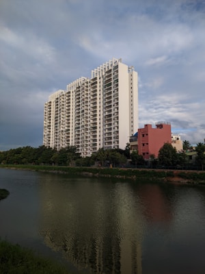 A tall, modern apartment building with multiple floors stands prominently near a body of water. The building is white with a sleek design and numerous balconies. In front of it, a smaller, reddish building is visible, along with lush green trees lining the water. The water reflects the apartment building and surrounding foliage. The sky is expansive and partly cloudy, with a serene blue hue.