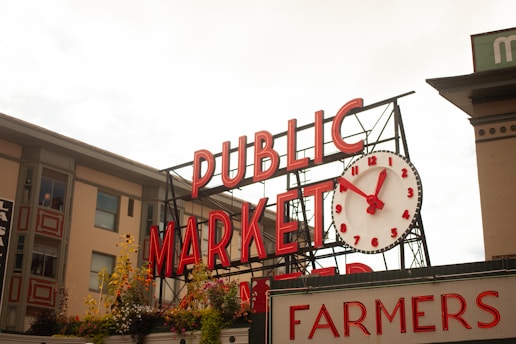 A prominent sign reads 'PUBLIC MARKET' with large red letters on a metal framework. Below the sign, a large white clock with red numbers and hands is visible. The sign is accompanied by another sign below that reads 'FARMERS' in red letters. There are various flower arrangements and plants in front of the signs and the building in the background has several windows with flower boxes.