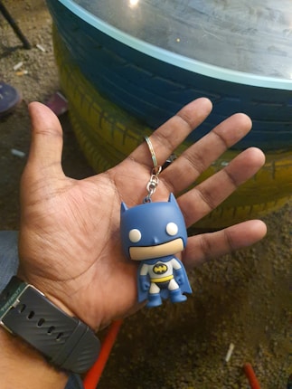 A hand holds a small keychain figure resembling a cartoonish superhero in a blue costume with a black bat symbol on its chest. The background has a round glass table and colorful painted tires, with the setting likely being outdoors.