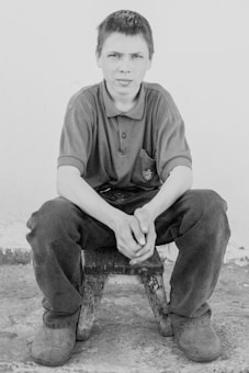 A young person is seated on a small wooden stool against a plain background, wearing a polo shirt and trousers. The expression on their face is neutral, with hands clasped loosely in front of them.
