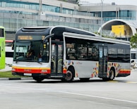 A public transit bus is captured in an urban environment, set against a modern building with a curved glass facade. The bus is white with red and orange accents and displays a route number and destination on its electronic display board.