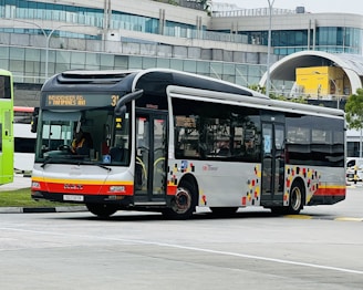 A public transit bus is captured in an urban environment, set against a modern building with a curved glass facade. The bus is white with red and orange accents and displays a route number and destination on its electronic display board.