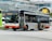 A public transit bus is captured in an urban environment, set against a modern building with a curved glass facade. The bus is white with red and orange accents and displays a route number and destination on its electronic display board.
