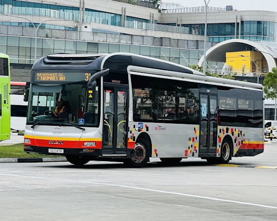 A public transit bus is captured in an urban environment, set against a modern building with a curved glass facade. The bus is white with red and orange accents and displays a route number and destination on its electronic display board.