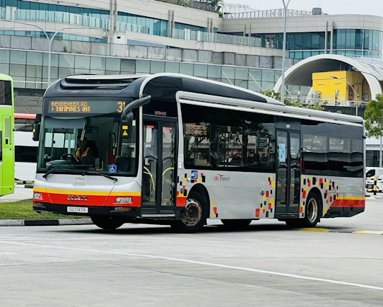 A public transit bus is captured in an urban environment, set against a modern building with a curved glass facade. The bus is white with red and orange accents and displays a route number and destination on its electronic display board.