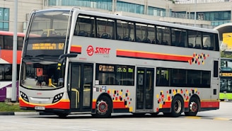 A double-decker bus with a modern design featuring a silver body with colorful geometric patterns on the sides. The bus is part of the SMRT fleet and displays route information on the front. It is stationed at what appears to be a bus terminal or parking area with buildings visible in the background.