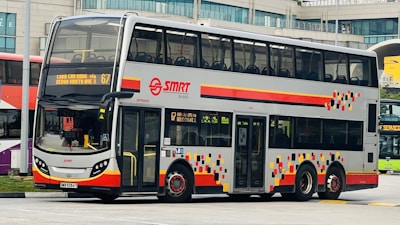 A double-decker bus with a modern design featuring a silver body with colorful geometric patterns on the sides. The bus is part of the SMRT fleet and displays route information on the front. It is stationed at what appears to be a bus terminal or parking area with buildings visible in the background.