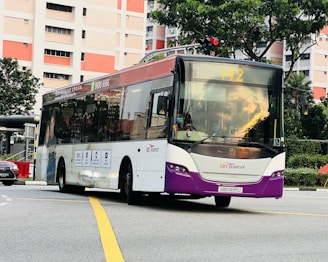 A public transit bus in motion on an urban road. The bus has a purple and white color scheme with the SBS Transit logo. It is displaying route number 142 and is surrounded by high-rise residential buildings and greenery.